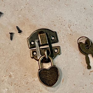 May include: A vintage-style metal clasp with a heart-shaped lock and two keys. The clasp is a dark brass color, with a rectangular base and a hinged top. Four small black screws are scattered around the clasp.