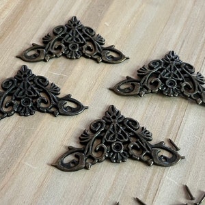 May include: Four ornate metal corner brackets in a triangular shape with a decorative floral design. The brackets are a dark bronze color and have a distressed finish. The brackets are shown on a light wood surface with small screws.
