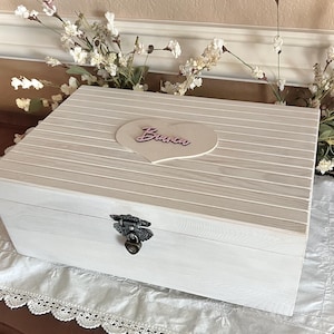 May include: A white wooden box with a light brown striped top and a heart-shaped accent that reads "Bianca". The box has a decorative metal clasp and is sitting on a white lace-trimmed cloth. Decorative flowers are in the background.
