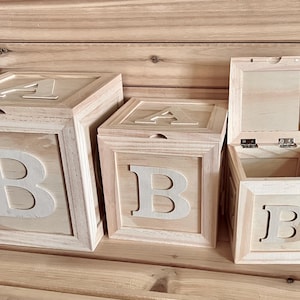 May include: Three light-colored wooden storage boxes shaped like alphabet blocks, arranged in descending size. Each box features a large white letter B on the front and a letter A on the top lid. The smallest box on the right has an open hinged lid.