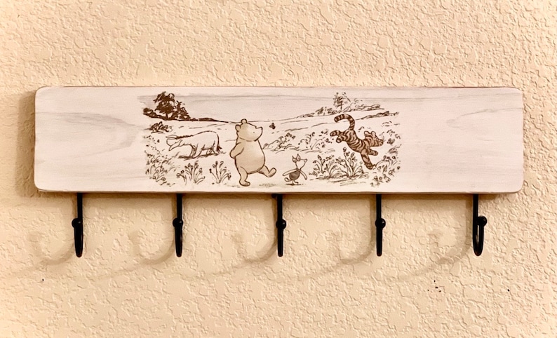 Op de afbeelding: Witte houten sleutelhanger met vijf haken en een Winnie de Poeh-thema. Het ontwerp toont Winnie de Poeh, Biggetje en Tijger die door een bos lopen.
