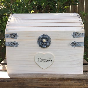 May include: A light-colored wooden chest with a curved top and metal hardware. The chest features a heart-shaped plaque with the name "Hannah" in script. The chest is designed with a vintage aesthetic.
