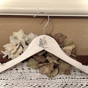 May include: A light-colored wooden clothes hanger with a silver hook. The hanger features a small illustration of a rabbit in a blue jacket. The hanger is decorated with white and brown flowers.