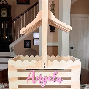 May include: A light-colored wooden basket with a scalloped edge and a handle, featuring a pink nameplate that reads "Angela." The basket has wheels and a built-in hanger rack.