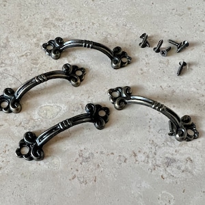 May include: Three antique-style metal drawer pulls with ornate details. Each pull has a curved shape and a decorative design at each end. The pulls are accompanied by eight small screws.