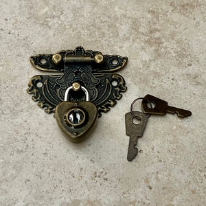 May include: Antique brass heart-shaped lock and key set with ornate details. The lock has a keyhole in the center of the heart and is attached to a decorative latch. The keys are small and have a simple design.
