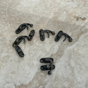 May include: Several antique bronze-colored metal latches are scattered on a light-colored, textured surface. The latches have a curved hook design and are attached to rectangular plates with screw holes, suitable for small boxes or chests.