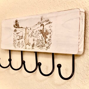 Op de afbeelding: Witte houten wandbevestigde sleutelhanger met een Winnie de Poeh en vrienden ontwerp. De hanger heeft vijf zwarte metalen haken.