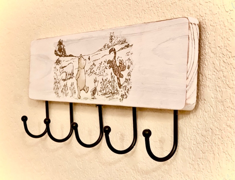 Op de afbeelding: Witte houten wandbevestigde sleutelhanger met een Winnie de Poeh en vrienden ontwerp. De hanger heeft vijf zwarte metalen haken.