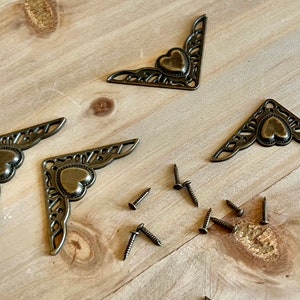 May include: Four antique gold corner brackets with a heart design and eight small screws. The brackets are ornate and have a filigree pattern.