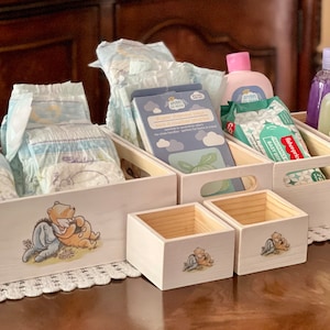 May include: Five white wooden crates with Winnie the Pooh designs. The crates are filled with baby supplies, including diapers, wipes, lotion, and baby wash.