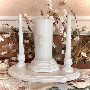 May include: A white oval wooden base with a beaded edge holds a central pillar candle featuring a small pearl-embellished cross. Two matching white taper candle holders stand on either side of the central pillar, each supporting a thin white candle.