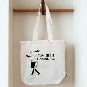Könnte beinhalten: Eine cremefarbene Tragetasche mit dem Text "Night Shift Social Club" und einer schwarzen Grafik einer Person, die Kaffeetassen trägt. Die Tasche hängt an einem Holzhaken.