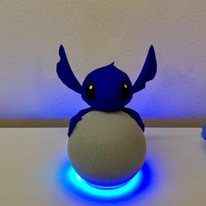 Può includere: Una statuetta blu di Stitch è posizionata sopra un altoparlante sferico grigio. La statuetta ha grandi occhi neri e orecchie. L'altoparlante poggia su una superficie bianca, illuminata da un anello blu brillante.