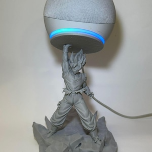 Può includere: Una statuetta grigia di un personaggio di Dragon Ball Z che sorregge un altoparlante intelligente. L'altoparlante è bianco con una parte superiore in rete grigia e una luce blu. La statuetta è su una base rocciosa.