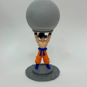Könnte beinhalten: Ein grauer, kugelförmiger Smart Speaker, der von einer Kunststofffigur eines muskulösen Charakters mit stacheligen schwarzen Haaren hochgehalten wird, der einen orangefarbenen Gi mit blauem Gürtel und blauen Stiefeln auf einem grauen Sockel trägt.