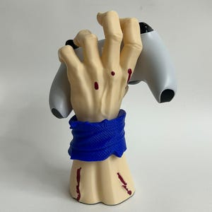 Può includere: Un supporto per controller a forma di mano color beige con schizzi rossi simili a sangue e un polsino blu testurizzato. La mano è in posizione di presa e tiene saldamente un controller di gioco bianco e nero tra le dita e il palmo.