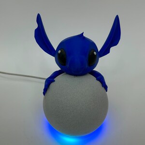 May include: A blue character figure with large ears and black eyes perched on top of a spherical gray smart speaker. The speaker features a glowing blue light ring at its base and a white power cord extending from the side of the device.