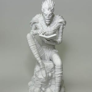 May include: A white figurine of a skeletal figure seated on a rock formation adorned with skulls. The figure is writing in a small book with a pen. The figure has spiky hair and is wearing a detailed outfit.