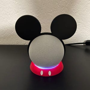 Könnte beinhalten: Ein intelligenter Lautsprecher in einem rot-schwarzen Mickey-Mouse-Ständer. Der Lautsprecher ist grau und hat unten ein weißes Licht. Der Ständer hat zwei weiße ovale Knöpfe und zwei schwarze Ohren.