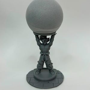 Könnte beinhalten: Ein grauer, kugelförmiger Smart Speaker auf einem grauen, 3D-gedruckten Ständer in Form einer muskulösen Anime-Figur mit stacheligen Haaren, die auf einem runden Sockel mit Steinmuster steht und den Lautsprecher mit beiden Armen hochhält.