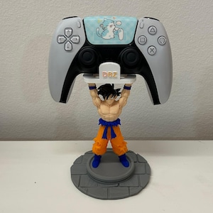 Può includere: Un supporto bianco per controller di videogiochi con una figura dai capelli neri, pantaloni arancioni e stivali blu che sostiene una piattaforma bianca con il testo DBZ. Il controller ha un adesivo azzurro con un gatto dei cartoni animati.