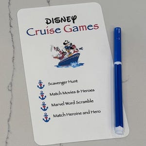 May include: A white card with the words "DISNEY Cruise Games" in blue and red. The card features a cartoon image of Disney characters on a boat. A blue marker is next to the card. The card lists four games: Scavenger Hunt, Match Movies & Heroes, Marvel Word Scramble, and Match Heroine and Hero.