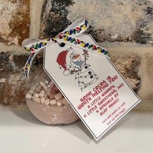 May include: A clear spherical ornament filled with hot cocoa mix and mini marshmallows, tied with a ribbon featuring colorful Christmas lights. A white tag attached shows a cartoon snowman wearing a Santa hat and the text: SOME COCOA IS WORTH MELTING FOR! A LITTLE WARMTH, A LITTLE SWEETNESS, AND A SPRINKLE OF OLAF MAGIC! Just add water or milk and sip the magic.