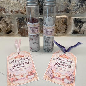 May include: Two clear glass tubes filled with bath salts, one with rose petals and one with lavender. Each tube has a label reading Soak your cares away princess. Two matching rectangular tags with pink ribbons read A royal soak for the princess in you.