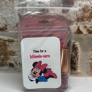 May include: A clear plastic bag containing a nail polish bottle and a pink pouch. A white card with the text "Time for a Minnie-cure" and a Minnie Mouse graphic is in front of the bottle. The nail polish is a glittery pink color.