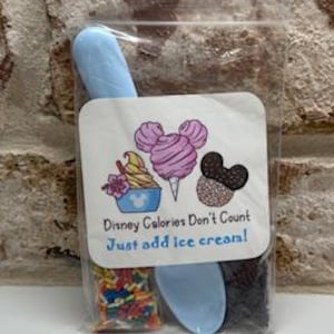 May include: A clear plastic bag containing a light blue ice cream scoop, sprinkles, and chocolate chips. A white label features a cartoon of a cotton candy cone, ice cream, and a chocolate cookie, with the text "Disney Calories Don't Count! Just add ice cream!"