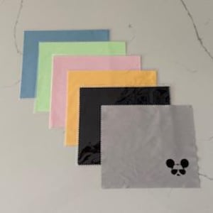 May include: A collection of six microfiber cleaning cloths in various colors: blue, green, pink, yellow, black, and gray. The gray cloth features a small black mouse face design in the corner. The cloths are arranged in a diagonal stack.