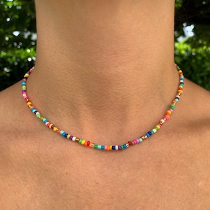 May include: A choker necklace made of small, round, multi-colored glass beads. The beads are arranged in a repeating pattern of various bright colors including red, orange, yellow, green, blue, purple, and pink, interspersed with metallic gold beads.