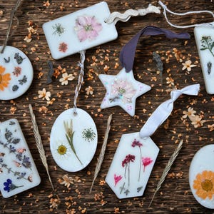 May include: A collection of white wax ornaments in diverse shapes, such as circles, rectangles, and a star, each featuring dried flowers. The ornaments are arranged on a rustic wooden surface, with natural embellishments.