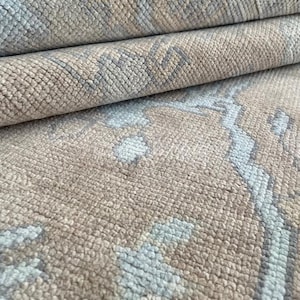 Puede incluir: Una alfombra tejida con una superficie texturizada y un patrón repetitivo en tonos beige claro y azul polvo suave. El material muestra una construcción de pelo denso y anudado con fibras visibles y un borde plegado.