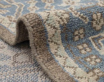 May include: Close-up of a rolled-up patterned rug. The rug displays a combination of light blue, beige, and grey colours. The edges are a darker shade of grey. The rug's design includes geometric and floral elements.