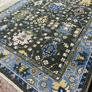 Puede incluir: Una alfombra verde oscuro con un patrón floral detallado en tonos de azul, amarillo y rosa. La alfombra tiene un borde azul claro con un diseño floral repetido y flecos en el borde. La alfombra es rectangular.