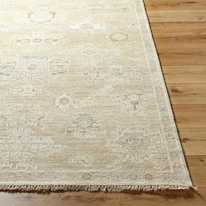Puede incluir: Una alfombra rectangular con una base de color beige claro y crema, que presenta un patrón repetitivo de motivos geométricos en azul y gris descoloridos. La alfombra tiene una superficie texturizada y flecos cortos en los bordes.