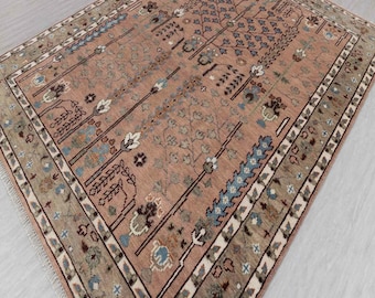 May include: A rectangular, hand-woven rug with a muted pink central field. The rug features a detailed pattern of stylized trees and floral motifs in shades of blue, brown, and white. The border is a mix of beige, brown, and white, adding a contrasting frame.