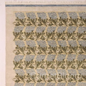 Puede incluir: Una alfombra rectangular con un patrón floral repetitivo de flores azul claro y tallos verde oscuro sobre un fondo beige. La alfombra tiene un borde azul fino cerca del borde superior y flecos cortos de color claro en la parte superior.