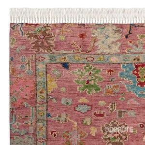 Puede incluir: Una alfombra rectangular con fondo rosa empolvado y un patrón floral y geométrico multicolor en tonos azul, verde, amarillo y marrón. La alfombra presenta un borde decorativo y flecos blancos en el borde superior.