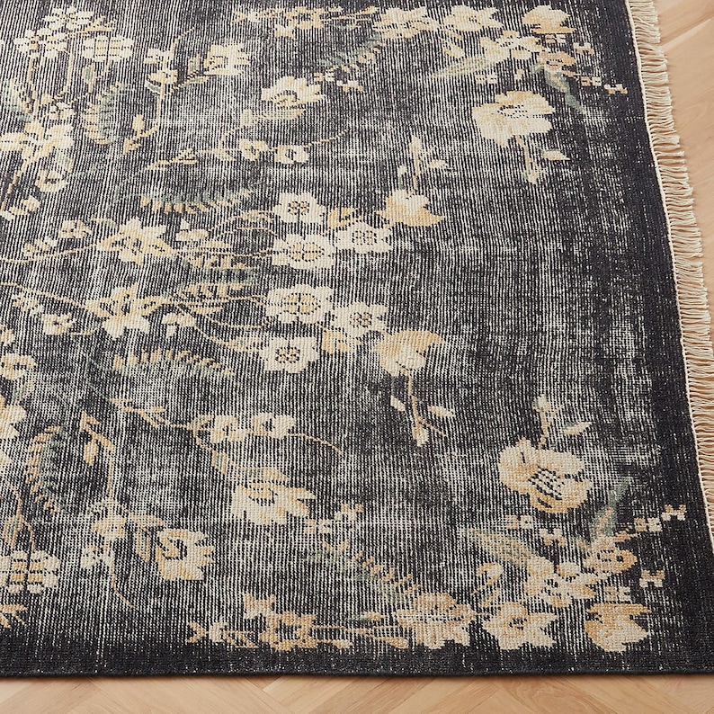 Puede incluir: Una alfombra de aspecto envejecido con un fondo gris oscuro y un estampado floral en crema y verde claro. La alfombra tiene un borde con flecos y una est&eacute;tica vintage, adecuada para salones o dormitorios.