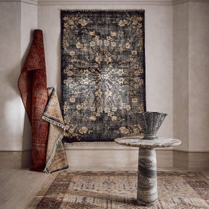 Puede incluir: Una habitaci&oacute;n con alfombras y una mesa de m&aacute;rmol. Una alfombra grande y oscura con un dise&ntilde;o floral cuelga de la pared. Tambi&eacute;n hay una alfombra roja enrollada y una alfombra m&aacute;s peque&ntilde;a con un borde beige. Una mesa de m&aacute;rmol con un jarr&oacute;n decorativo est&aacute; delante.