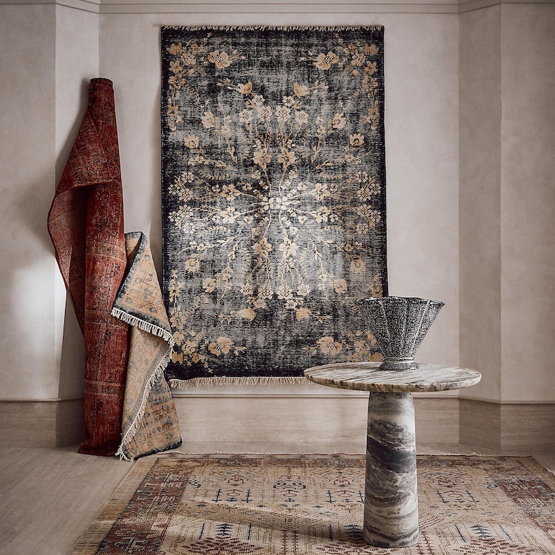 Puede incluir: Una habitaci&oacute;n con alfombras y una mesa de m&aacute;rmol. Una alfombra grande y oscura con un dise&ntilde;o floral cuelga de la pared. Tambi&eacute;n hay una alfombra roja enrollada y una alfombra m&aacute;s peque&ntilde;a con un borde beige. Una mesa de m&aacute;rmol con un jarr&oacute;n decorativo est&aacute; delante.