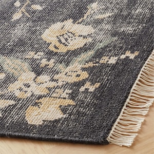 Puede incluir: Primer plano de una alfombra gris oscuro con un estampado floral en crema y amarillo. La alfombra tiene un aspecto desgastado y un borde con flecos. La alfombra est&aacute; sobre un suelo de madera.