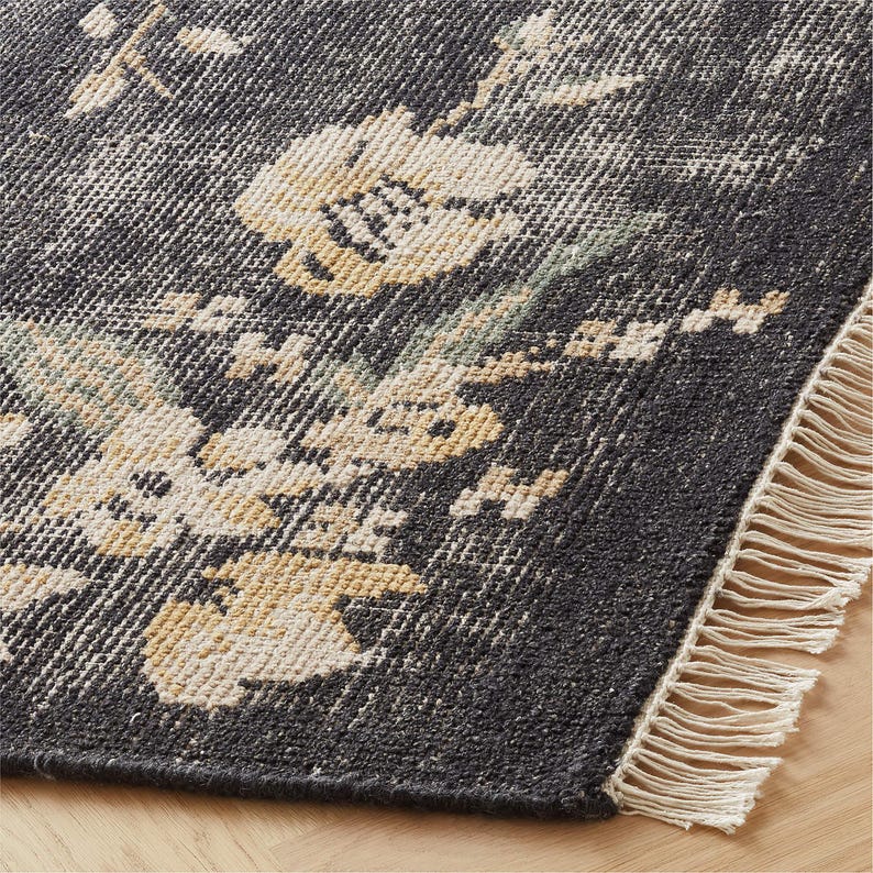 Puede incluir: Primer plano de una alfombra gris oscuro con un estampado floral en crema y amarillo. La alfombra tiene un aspecto desgastado y un borde con flecos. La alfombra est&aacute; sobre un suelo de madera.