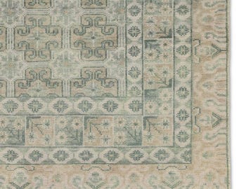 May include: A section of a rectangular rug with a detailed, geometric pattern. The rug features a combination of light blue, beige, and tan colours. The design includes repeating shapes and borders, creating a textured appearance. The rug's edge is visible, showing a fringed border.
