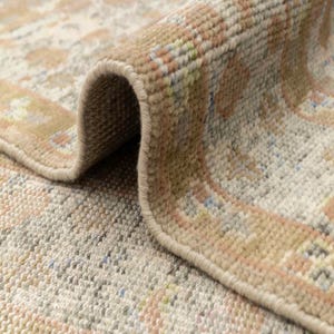 May include: Close-up of a rolled-up area rug with a detailed, textured pattern. The rug features a beige and cream background with accents of peach, blue, and green. The edges are neatly finished with a light brown border, showcasing the rug's intricate design.