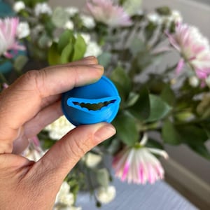 Peut inclure: Un objet circulaire bleu vif avec un motif de bouche souriante incurvée. L'objet est tenu dans une main, avec un arrangement floral flou en arrière-plan. Il semble s'agir d'un objet décoratif ou d'un petit récipient.