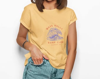May include: Yellow t-shirt with a graphic of a wave and the text "WAVE BULLY SURF CLUB CALIFORNIA, USA". The model is wearing blue denim shorts and yellow-tinted glasses. The shirt is made of a soft material.